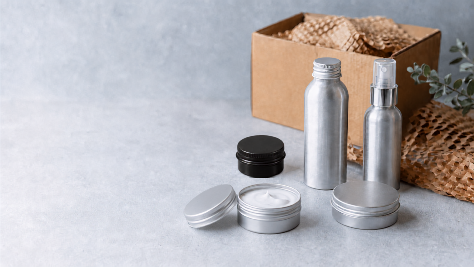 Gamme d’emballages cosmétiques en aluminium, comprenant des flacons, un vaporisateur et des pots, présentée avec une boîte en carton et du papier de calage.