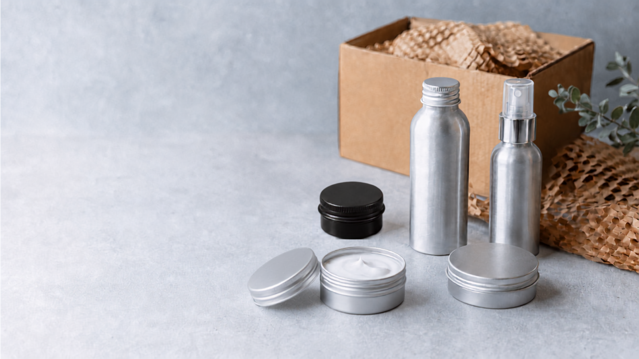 Gamme d’emballages cosmétiques en aluminium, comprenant des flacons, un vaporisateur et des pots, présentée avec une boîte en carton et du papier de calage.