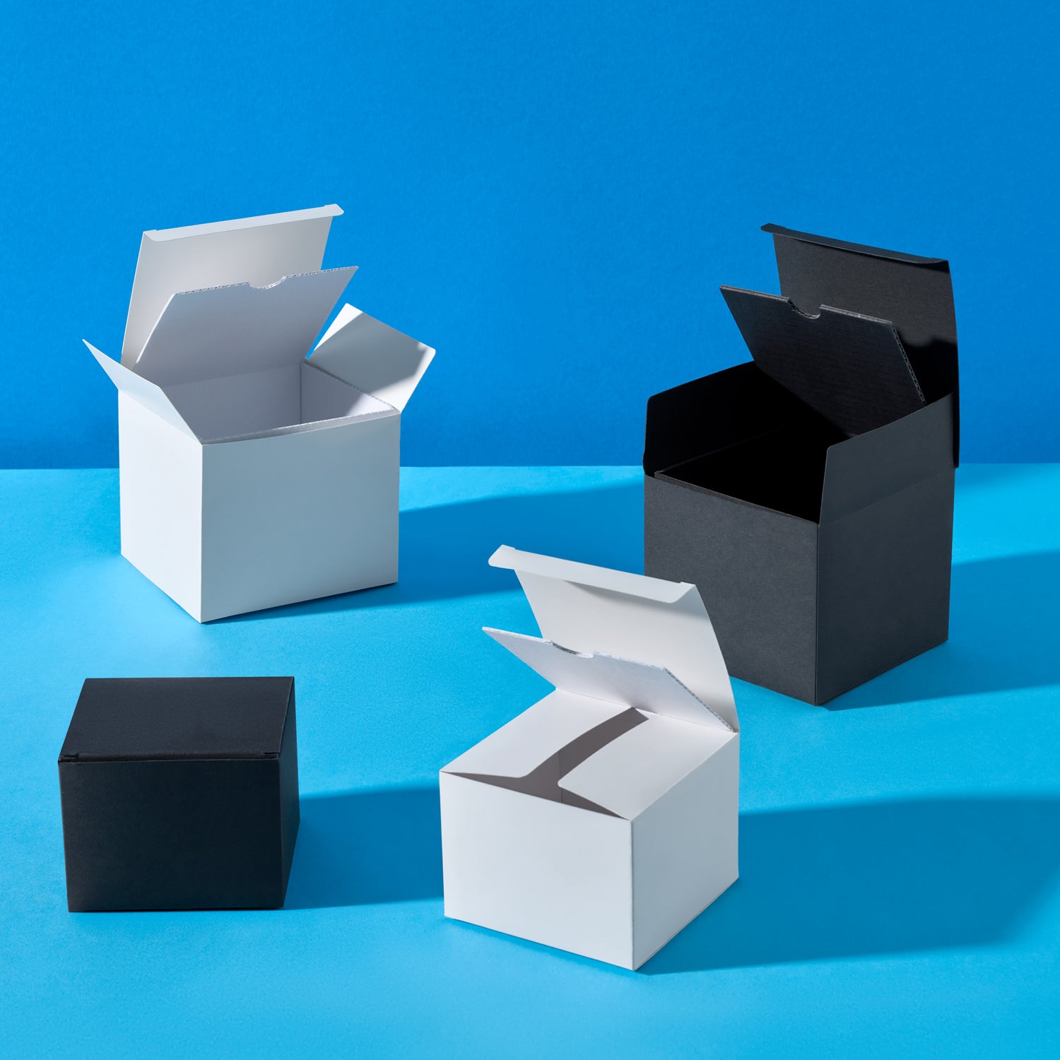 Plusieurs boîtes en carton noir et blanc, ouvertes et fermées, sont disposées sur un fond bleu. De forme carrée, les boîtes présentent des rabats surélevés, illustrant différents angles et positions d'ouverture. L'éclairage met en valeur la texture lisse du carton et le contraste entre les deux couleurs.